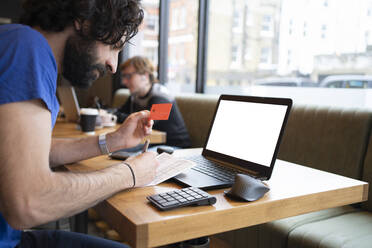 Männlicher Unternehmer, der einen Kredit in der Hand hält, während er eine Online-Zahlung am Laptop im Büro vornimmt - FBAF01688