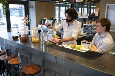 Auszubildender sieht männlichen Barkeeper an, der Früchte in ein Glas an der Theke legt - FBAF01741