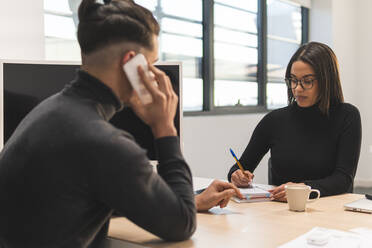 Geschäftsmann, der mit einem Smartphone spricht, während eine Kollegin im Büro in ihr Tagebuch schreibt - JAQF00427