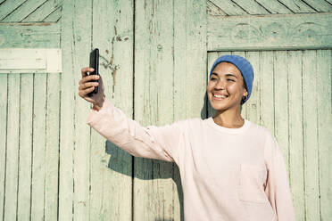 Glückliche Frau mit Strickmütze nimmt Selfie durch Handy während sonnigen Tag - RCPF00921