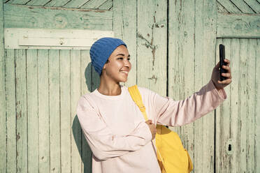 Glückliche Frau mit Strickmütze, die einen Rucksack trägt, während sie ein Selfie mit ihrem Mobiltelefon vor der Tür macht - RCPF00923