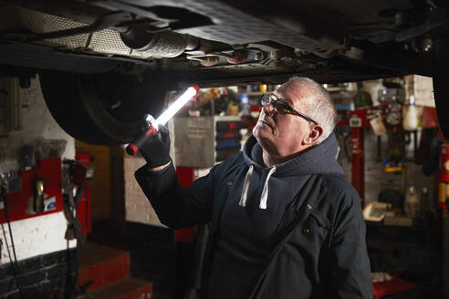 Älterer männlicher Mechaniker mit Taschenlampe, der ein Auto in einer Werkstatt untersucht - PMF01742