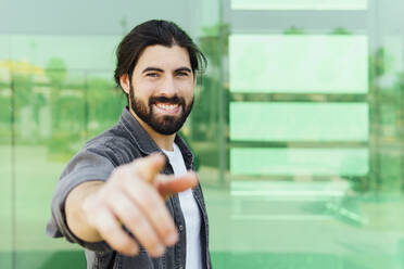 Smiling young man gesturing while standing in front of glass wall - MIMFF00637