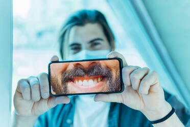 Junger Mann zeigt zahniges Lächeln auf dem Handy während des Covid-19 - MIMFF00643