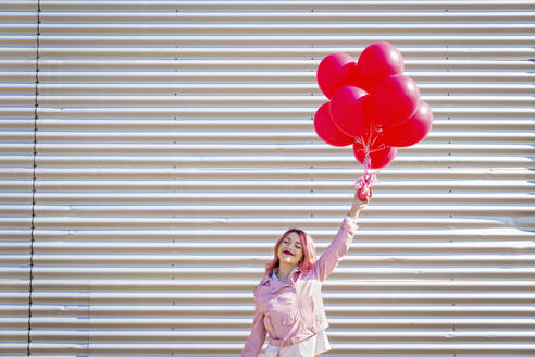 Happy woman with eyes closed holding bunch on balloons in front of metal wall - EIF00716