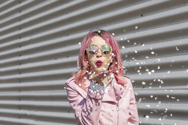 Junge Frau mit rosa Haaren bläst Papierkonfetti vor einer Wellblechwand - EIF00723