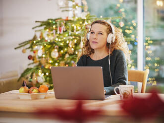Frau mit Kopfhörern, die wegschaut, während sie an Weihnachten vor einem Laptop sitzt - PWF00274