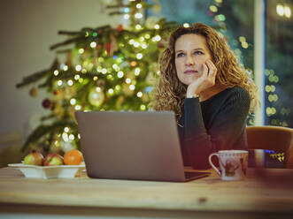 Frau träumt, während sie zu Hause vor ihrem Laptop sitzt, während der Weihnachtszeit - PWF00275