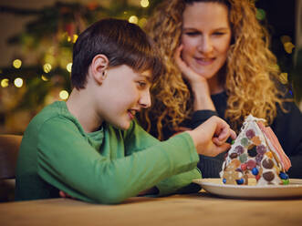Lächelnder Junge beim Verzieren eines Lebkuchenhauses durch seine Mutter während der Weihnachtszeit - PWF00278
