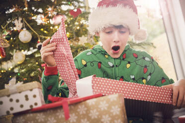 Aufgeregter Junge mit Weihnachtsmannmütze packt zu Hause ein Geschenk mit Weihnachtsbaum im Hintergrund aus - PWF00283
