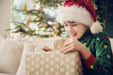 Niedlicher Junge mit Weihnachtsmannmütze packt an Weihnachten zu Hause ein Geschenk aus - PWF00285