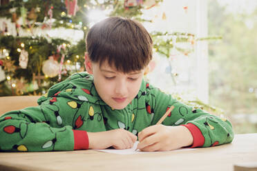 Cute boy writing on paper at table with Christmas tree in background at home - PWF00287