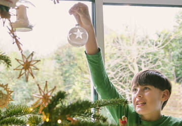 Cute boy decorating Christmas tree by window at home - PWF00295