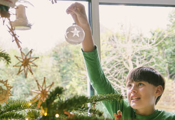 Niedlicher Junge schmückt Weihnachtsbaum am Fenster zu Hause - PWF00295