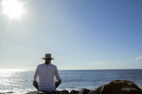 Mature man sitting on rock in front of sea during sunny day - PSTF00893