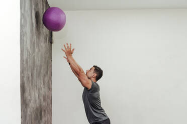 Mittlerer Erwachsener, männlicher Athlet, der mit einem Medizinball an einer Wand im Fitnessstudio trainiert - EBBF02871