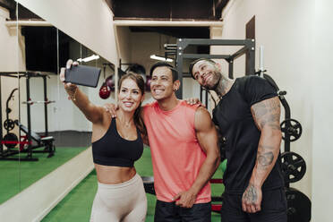 Lächelnde Sportlerin, die ein Selfie mit ihren männlichen Freunden im Fitnessstudio macht - EBBF02875