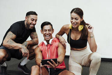 Laughing sportsman using mobile phone with friends around in gym - EBBF02881