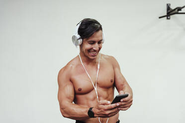 Hemdloser Sportler mit Kopfhörern, der lächelnd sein Handy im Fitnessstudio benutzt - EBBF02883