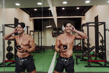 Lächelnder mittelgroßer Erwachsener, der Kopfhörer einstellt, während er am Spiegel im Fitnessstudio steht - EBBF02884