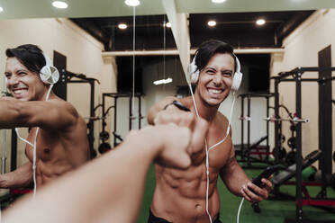 Fröhlicher Sportler mit Kopfhörern und Mobiltelefon, der einem Freund im Fitnessstudio die Faust gibt - EBBF02886