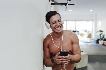 Shirtless male sportsperson with headphones and mobile phone leaning on white wall at health club - EBBF02888