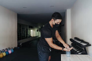 Sportsperson wearing protective face mask using hand sanitizer in health club - EBBF02894