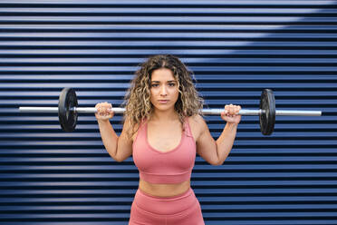 Young female athlete standing with barbell in front of blue wall - AODF00391