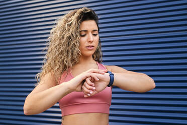 Schöne Sportlerin, die die Zeit auf ihrer Armbanduhr an der blauen Wand kontrolliert - AODF00394