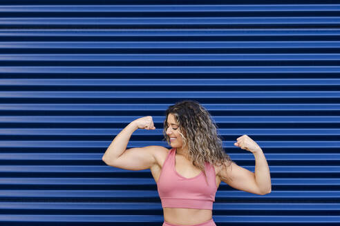 Glückliche Sportlerin beim Muskeltraining vor einer blauen Wand - AODF00417