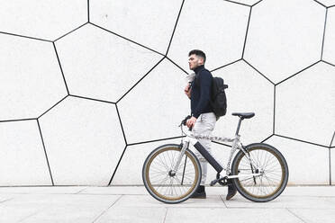 Young man wheeling bicycle by textured white wall - JAQF00439