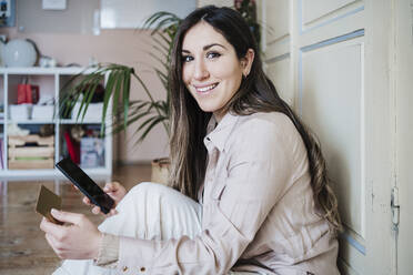 Cheerful woman holding smart phone and credit card while sitting at home - EBBF02896