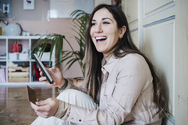 Laughing woman holding credit card and smart phone while sitting at home - EBBF02897