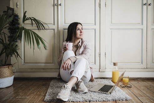 Thoughtful businesswoman looking away while sitting on rug at home - EBBF02904