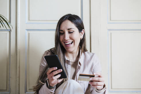 Happy woman using smart phone while holding credit card at home - EBBF02919