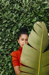 Woman looking through banana leaf while standing in front of green plants - TCEF01731