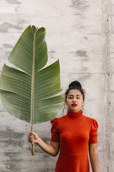 Young woman in turtleneck red dress holding green banana leaf in front of wall - TCEF01732