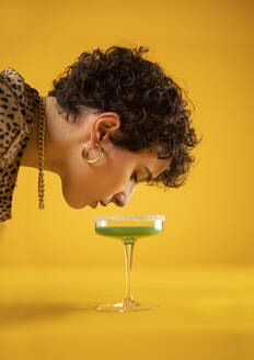 Young woman looking at cocktail in glass on yellow background - AXHF00238