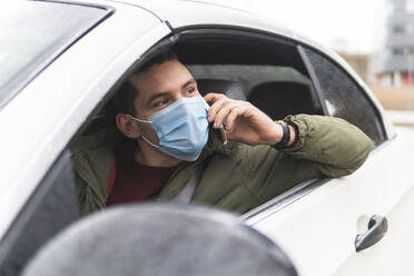 Young man wearing protective face mask talking on mobile phone while sitting in car - JAQF00441