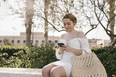 Young woman in white dress using mobile phone while sitting at park - EGAF02188