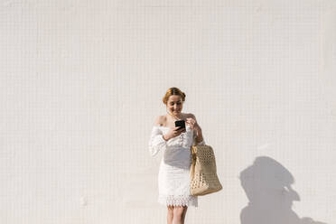 Smiling woman in white dress using mobile phone in front of white wall - EGAF02189