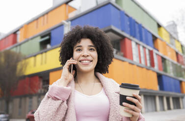 Smiling curly hair woman with disposable cup talking on mobile phone - JCCMF01588