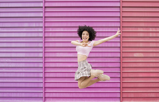 Carefree woman showing peace sign while jumping against multi colored wall - JCCMF01592