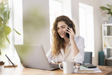 Zufriedene Unternehmerin, die mit ihrem Smartphone telefoniert, während sie einen Laptop am Schreibtisch im Heimbüro benutzt - SBOF03592