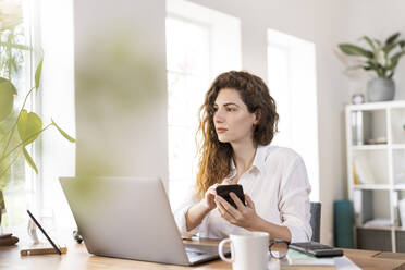 Beautiful female professional looking away while holding mobile phone at desk - SBOF03593