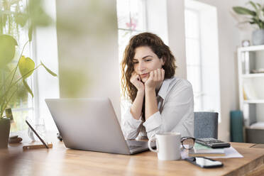 Businesswoman with hand on chin looking at laptop in home office - SBOF03594