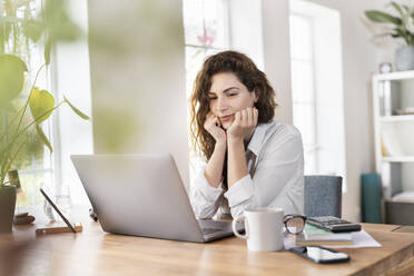 Geschäftsfrau mit Hand am Kinn, die auf einen Laptop im Heimbüro schaut - SBOF03594