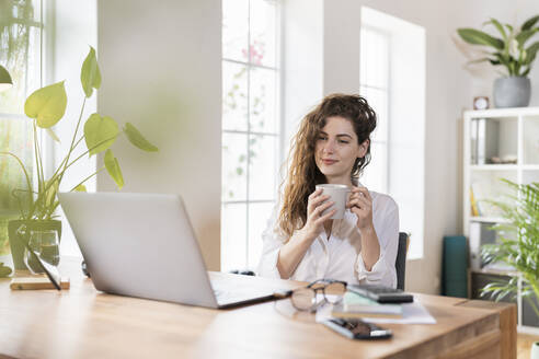 Schöne Freiberuflerin trinkt Kaffee und schaut auf den Laptop im Home Office - SBOF03595