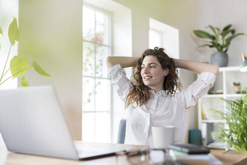 Lächelnde weibliche Fachkraft mit Händen hinter dem Kopf, die sich am Schreibtisch im Heimbüro entspannt - SBOF03597