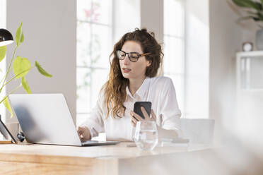 Female professional with eyeglasses working on laptop while holding smart phone at desk - SBOF03599